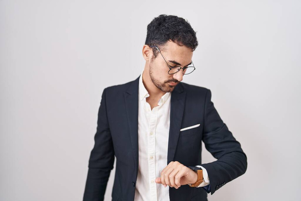 Handsome business hispanic man standing over white background checking the time on wrist watch, relaxed and confident  - Photo, Image