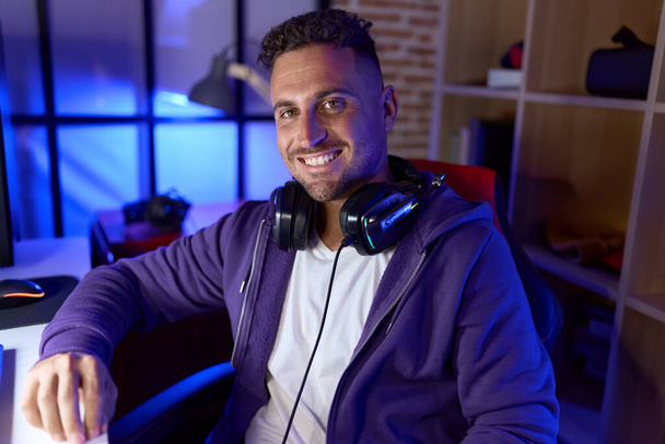 Молодий іспаномовний чоловік стример посміхається впевнено сидячи на столі в ігровій кімнаті
 - Фото, зображення