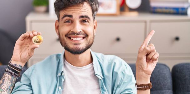 Young hispanic man with tattoos holding cardano cryptocurrency coin smiling happy pointing with hand and finger to the side  - Foto, afbeelding