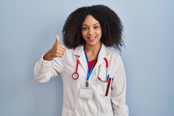 Giovane donna afroamericana vestita da medico uniforme e stetoscopio fare pollici felici in alto gesto con la mano. approvazione espressione guardando la fotocamera che mostra il successo.  - Foto, immagini