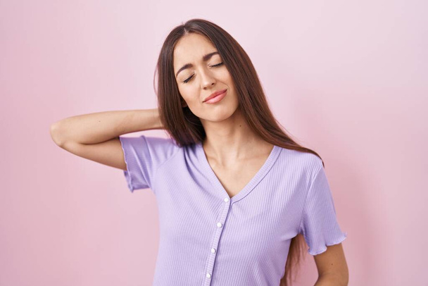 Mujer hispana joven con el pelo largo de pie sobre fondo rosa sufriendo de dolor de cuello lesión, tocando el cuello con la mano, dolor muscular  - Foto, Imagen