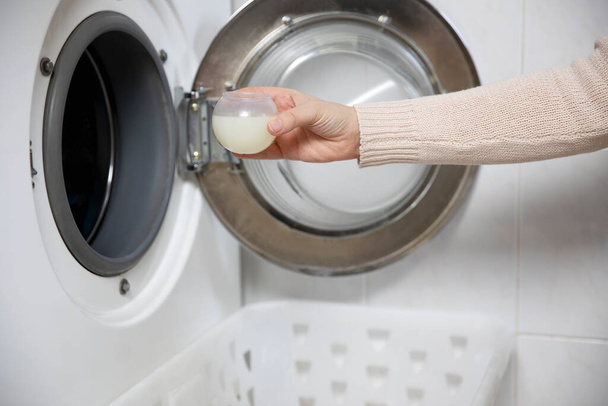 Ženské ruce nalévají tekutý prací prášek do šálku na míru. Pračka a oblečení v bílém moderním pokoji zblízka - Fotografie, Obrázek