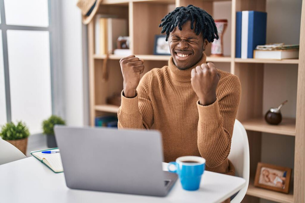 コンピュータのラップトップを使用して働いている恐ろしいロックを持つ若いアフリカの男は非常に満足し、腕を上げ、笑顔と成功のために叫んで勝者のジェスチャーを行うことに興奮しています。お祝いのコンセプト.  - 写真・画像
