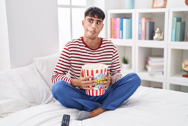 Giovane uomo ispanico mangiare popcorn seduto sul letto a guardare un film facendo faccia di pesce con la bocca e occhi strabici, pazzo e comico.  - Foto, immagini