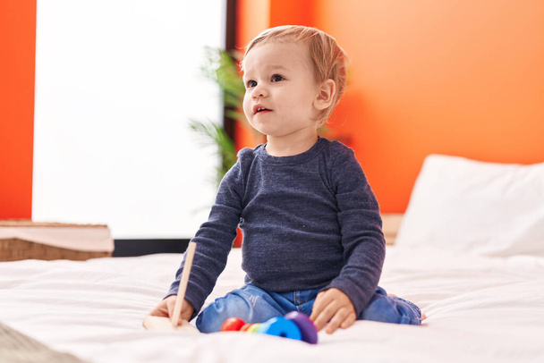 Schattige blonde peuter spelen met hoepels speelgoed zitten op bed in de slaapkamer - Foto, afbeelding