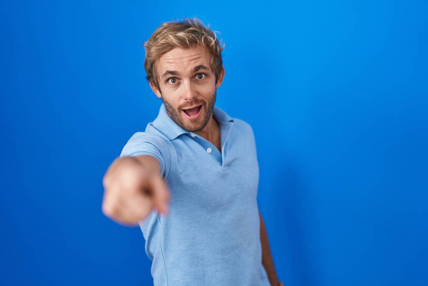 Blanke man die over een blauwe achtergrond staat en ontevreden en gefrustreerd naar de camera wijst, boos en woedend op jou  - Foto, afbeelding