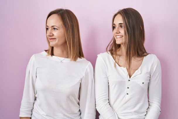 Middle age mother and young daughter standing over pink background looking away to side with smile on face, natural expression. laughing confident.  - Photo, Image