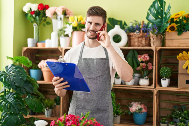 Young caucasian man florist talking on smartphone reading document at flower shop - Photo, Image