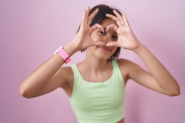 Jeune fille debout sur fond rose faisant forme de coeur avec la main et les doigts souriant en regardant à travers signe  - Photo, image