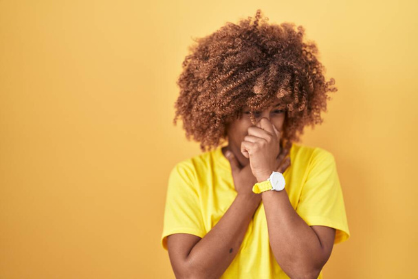 Young hispanic woman with curly hair standing over yellow background smelling something stinky and disgusting, intolerable smell, holding breath with fingers on nose. bad smell  - Photo, Image