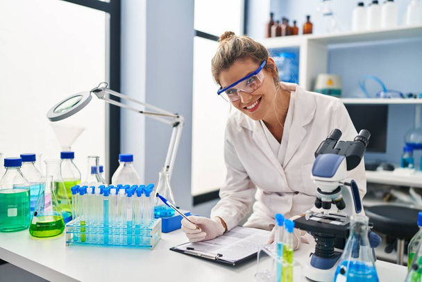 Młoda blondynka w mundurze naukowca pisząca na tablicy w laboratorium - Zdjęcie, obraz