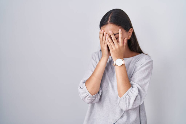 Молода іспаномовна жінка стоїть над білим тлом з сумним виразом, що покриває обличчя руками під час плачу. концепція депресії
.  - Фото, зображення