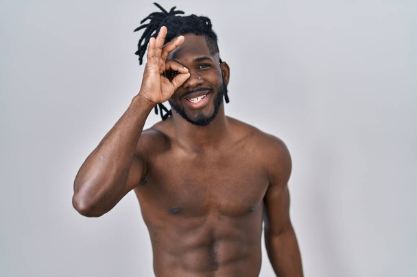 Африканский мужчина с дредами стоит без рубашки на изолированном фоне улыбаясь счастливо делает хорошо знак с рукой на глазу глядя сквозь пальцы  - Фото, изображение