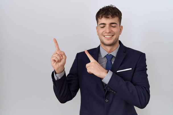 Junger hispanischer Geschäftsmann in Anzug und Krawatte lächelt lächelnd in die Kamera und zeigt mit zwei Händen und Fingern zur Seite.  - Foto, Bild