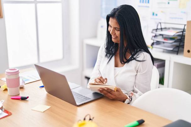 Middelbare leeftijd Spaanse vrouw zakenman met behulp van laptop schrijven op notebook op kantoor - Foto, afbeelding