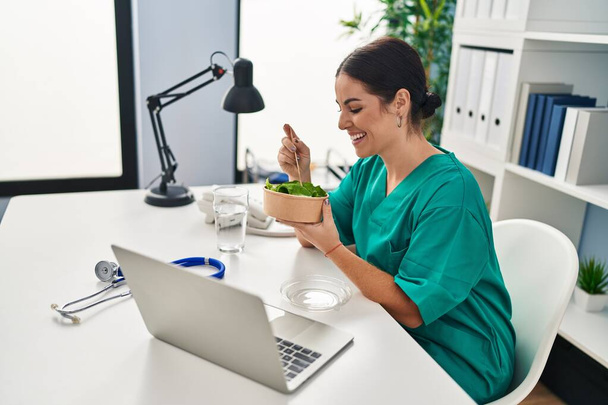 Young beautiful hispanic woman doctor smiling confident eating salad at clinic - 写真・画像