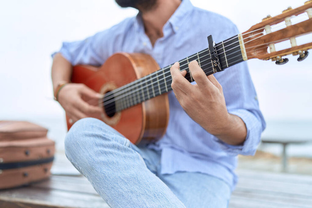 Young hispanic man musician playing classical guitar sitting on bench at seaside - Fotoğraf, Görsel