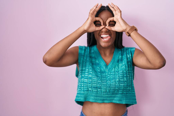 Joven afroamericano con trenzas paradas sobre fondo rosa haciendo un gesto aceptable como binoculares sacando la lengua, ojos mirando a través de los dedos. expresión loca.  - Foto, imagen
