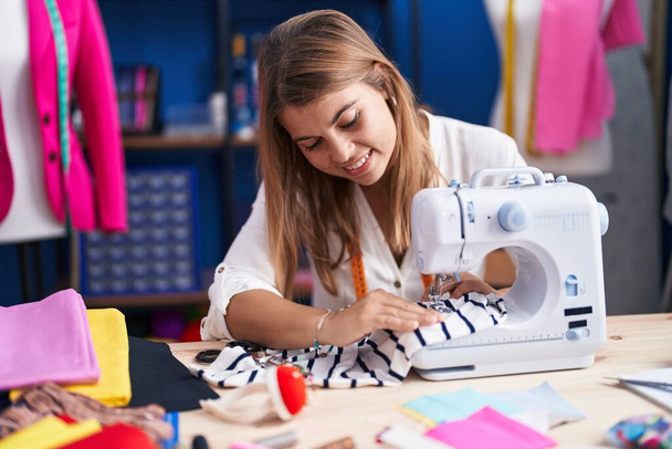 Jonge vrouw kleermaker glimlachen zelfverzekerd met behulp van naaimachine in naaiatelier - Foto, afbeelding