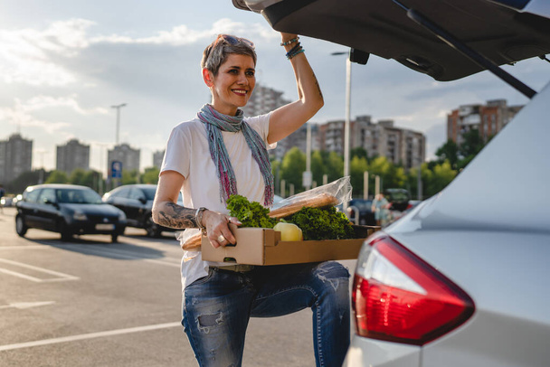 One woman mature caucasian female standing by the back trunk of her car on the parking lot of the supermarket shopping mall or grocery store with vegetables food in box putting them in the vehicle - Foto, immagini