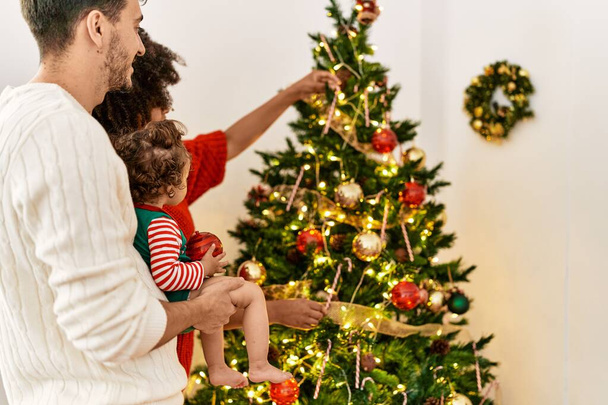 Pareja e hija sonriendo confiadas decorando el árbol de Navidad en casa - Foto, imagen