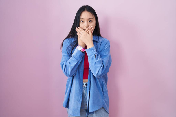 Mladá Asiatka, stojící nad růžovým pozadím, šokovaně zakryla ústa rukama, aby nedošlo k omylu. tajný koncept.  - Fotografie, Obrázek