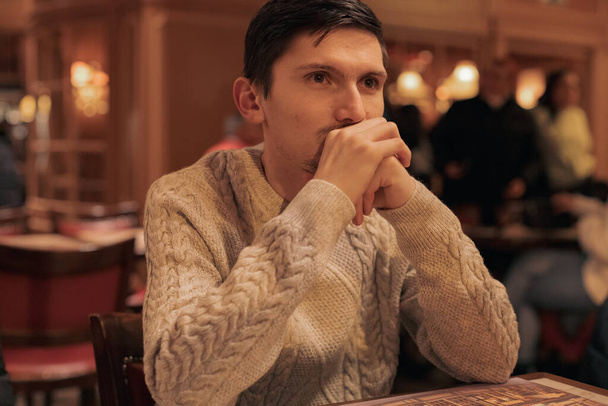 Portrait of one young handsome caucasian brunnet guy in a knitted sweater sitting thoughtfully with his chin propped up in his hands at a table in a restaurant, close-up side view. Concept lifestyle of men, real emotions. - Foto, Imagen