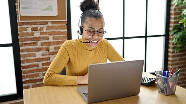 若いアフリカ系アメリカ人女性のビジネスワーカーは、オフィスでヘッドセットを着用したラップトップを使用して - 写真・画像
