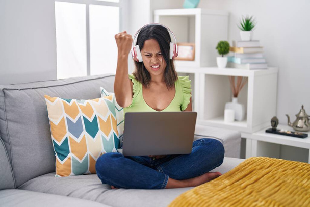 Hispanic young woman using laptop at home angry and mad raising fist frustrated and furious while shouting with anger. rage and aggressive concept.  - Foto, Imagem