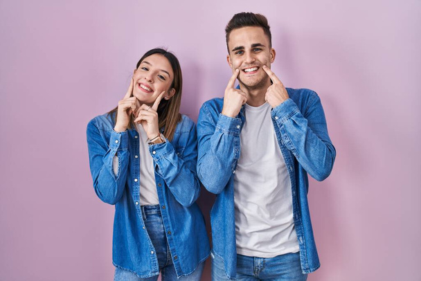 Νεαρό ζευγάρι ισπανόφωνων στέκεται πάνω από ροζ φόντο χαμογελώντας με ανοιχτό στόμα, δάχτυλα δείχνοντας και αναγκάζοντας χαρούμενο χαμόγελο  - Φωτογραφία, εικόνα