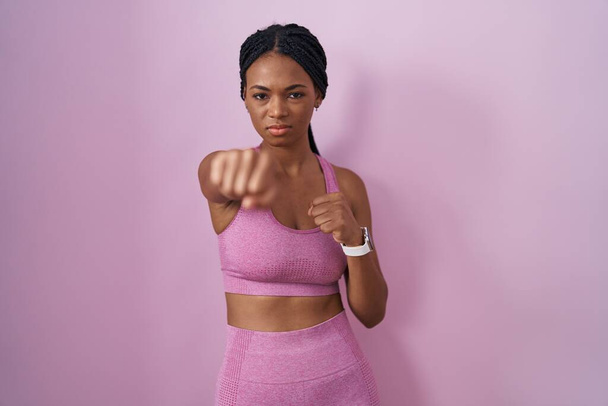 Αφροαμερικανή γυναίκα με πλεξούδες φορώντας αθλητικά ρούχα πάνω από ροζ φόντο γροθιά γροθιά για την καταπολέμηση, επιθετική και θυμωμένη επίθεση, απειλή και βία  - Φωτογραφία, εικόνα