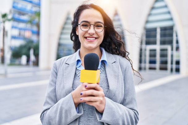 Νεαρή μελαχρινή γυναίκα κρατώντας μικρόφωνο δημοσιογράφος αναζητούν θετική και ευτυχισμένη στέκεται και χαμογελά με αυτοπεποίθηση χαμόγελο δείχνει τα δόντια  - Φωτογραφία, εικόνα