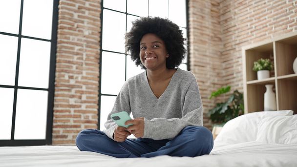 African american woman using smartphone sitting on bed at bedroom - Fotó, kép