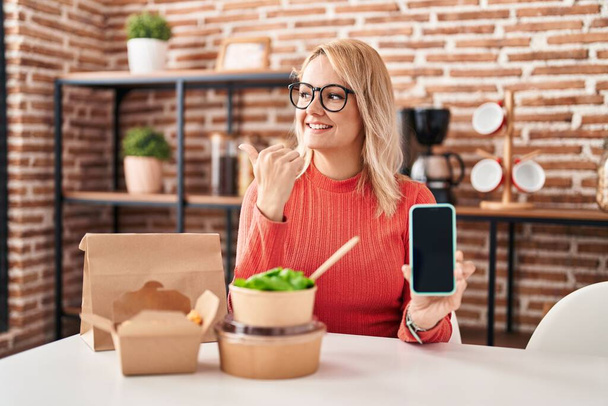 Blondýny žena jíst odnést jídlo ukazující smartphone obrazovku ukazující palec nahoru na stranu usmívat šťastný s otevřenými ústy  - Fotografie, Obrázek