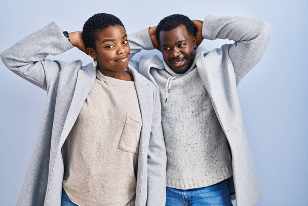 Молодая африканская американская пара, стоящая на синем фоне вместе расслабляющая и растягивающая, руки и руки за головой и шеей улыбаются счастливо  - Фото, изображение