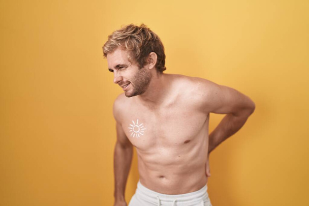 Белый мужчина, стоящий без рубашки с солнцезащитным кремом, страдающий от боли в спине, касаясь спины рукой, мышечная боль  - Фото, изображение