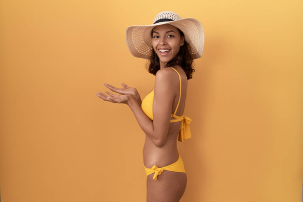 Mulher hispânica jovem usando biquíni e chapéu de verão apontando para o lado com as mãos abertas palmas mostrando espaço de cópia, apresentando propaganda sorrindo feliz animado  - Foto, Imagem