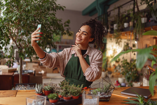 Χαμογελαστή γυναίκα ανθοπώλης φωτογραφίζει με τα φυτά της στο δικό της ανθοπωλείο στούντιο. Θολή φόντο - Φωτογραφία, εικόνα