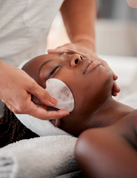 Black woman face, relax and gua sha stone spa treatment of a young female with facial. Skincare, beauty and wellness clinic with client feeling calm and zen from massage and dermatology glow. - Photo, image