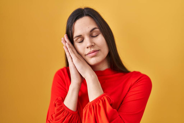 Junge hispanische Frau steht vor gelbem Hintergrund und schläft müde träumend und posiert mit den Händen zusammen, während sie mit geschlossenen Augen lächelt.  - Foto, Bild