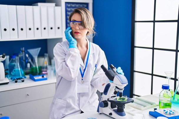Young woman scientist using microscope talking on smartphone at laboratory - Фото, изображение