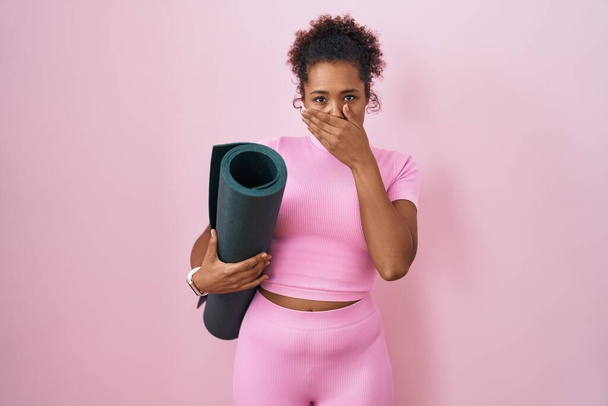 Mujer hispana joven con el pelo rizado sosteniendo estera de yoga sobre fondo rosa impactó cubriendo la boca con las manos por error. concepto secreto.  - Foto, imagen