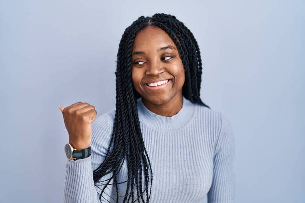 African american woman standing over blue background smiling with happy face looking and pointing to the side with thumb up.  - Photo, Image