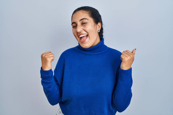 Young hispanic woman standing over isolated background very happy and excited doing winner gesture with arms raised, smiling and screaming for success. celebration concept.  - Foto, imagen