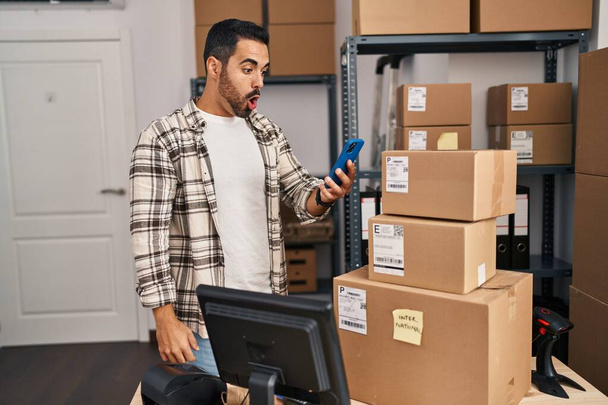 Young hispanic man with beard working at small business ecommerce with smartphone scared and amazed with open mouth for surprise, disbelief face  - Photo, Image