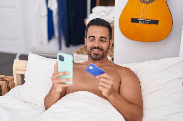 Jeune homme hispanique utilisant smartphone et carte de crédit couché sur le lit dans la chambre - Photo, image