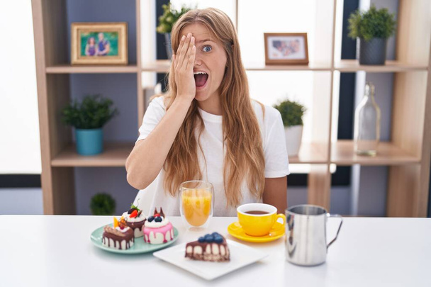 Young caucasian woman eating pastries t for breakfast covering one eye with hand, confident smile on face and surprise emotion.  - Photo, Image