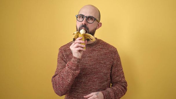 Fiatal kopasz férfi eszik banán felett elszigetelt sárga háttér - Fotó, kép