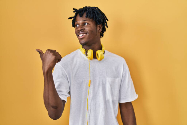 Jeune homme africain avec dreadlocks debout sur fond jaune souriant avec le visage heureux regardant et pointant vers le côté avec le pouce vers le haut.  - Photo, image