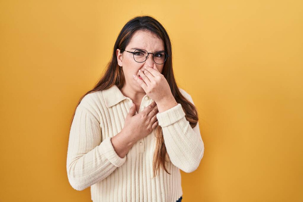 Young hispanic woman standing over yellow background smelling something stinky and disgusting, intolerable smell, holding breath with fingers on nose. bad smell  - Foto, imagen